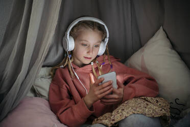 Mädchen mit Kopfhörern und Mobiltelefon auf dem Vordach zu Hause - NJAF00841