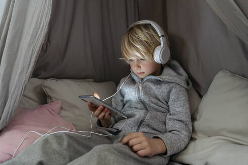 Junge mit Kopfhörern, der sein Smartphone zu Hause auf dem Vordach benutzt - NJAF00844