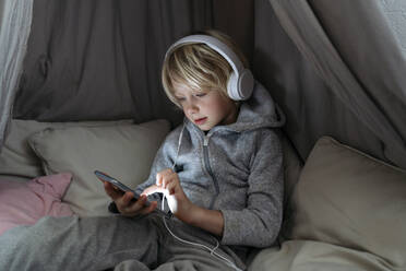 Boy wearing headphones using internet on smart phone in canopy at home - NJAF00846