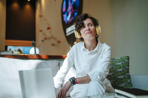 Nachdenkliche Geschäftsfrau, die mit einem Laptop in der Hotellobby sitzt und über drahtlose Kopfhörer Musik hört - MDOF01893