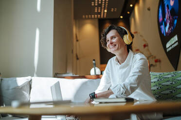 Smiling thoughtful businesswoman listening to music through wireless headphones sitting in hotel lobby - MDOF01895