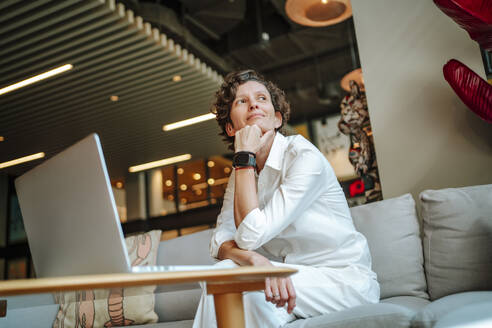 Nachdenkliche Geschäftsfrau sitzt mit der Hand am Kinn neben dem Laptop in der Hotellobby - MDOF01897