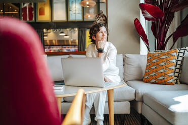 Nachdenkliche Geschäftsfrau sitzt mit der Hand am Kinn neben ihrem Laptop in der Lobby eines Hotels - MDOF01898