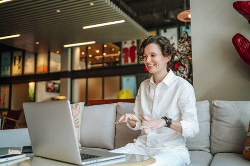 Happy businesswoman gesturing on video call through laptop in hotel lobby - MDOF01902