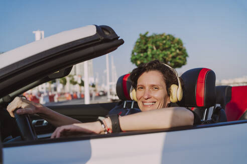 Lächelnde Frau mit drahtlosen Kopfhörern im Cabrio an einem sonnigen Tag - MDOF01928