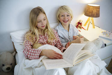 Glückliche Geschwister, die zu Hause auf dem Bett sitzend ein Buch lesen - NJAF00852