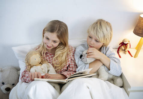 Lächelndes Mädchen, das mit seinem Bruder auf dem Bett sitzend zu Hause ein Buch liest - NJAF00853