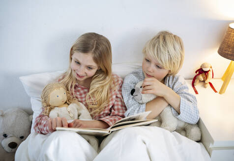 Smiling girl reading book with brother sitting on bed at home - NJAF00853