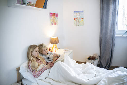 Blond brother and sister reading book on bed at home - NJAF00854