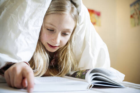 Blond covered covered in blanket reading book at home - NJAF00855
