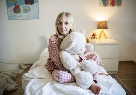 Smiling girl in pajamas sitting with teddy bear on bed at home - NJAF00857