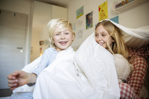 Happy brother and sister playing with blanket on bed at home - NJAF00859