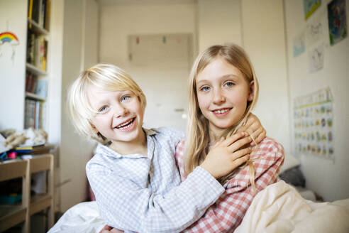 Smiling boy sitting with arm around sister in bedroom at home - NJAF00862