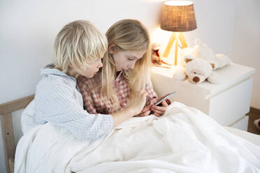 Neugierige Geschwister benutzen ihr Smartphone, während sie zu Hause auf dem Bett sitzen - NJAF00873