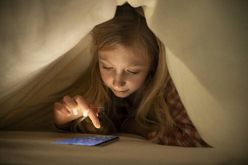 Mädchen benutzt Smartphone unter der Decke auf dem Bett zu Hause - NJAF00878
