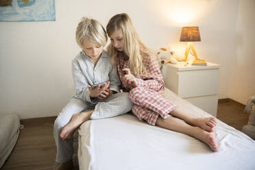Geschwister im Schlafanzug mit Smartphone auf dem heimischen Bett sitzend - NJAF00879