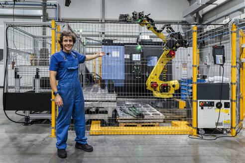 Smiling maintenance engineer gesturing at robotic arm in modern factory - AAZF01714