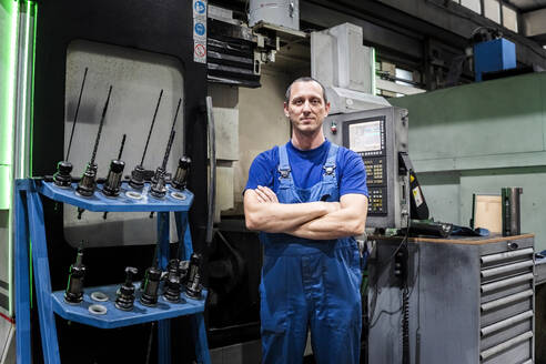 Selbstbewusster reifer Techniker in Uniform, der mit verschränkten Armen vor einer Maschine in der Industrie steht - AAZF01720