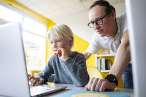 Blonder Junge, der einen Laptop benutzt, während sich der Professor im Klassenzimmer an den Schreibtisch lehnt - NJAF00915