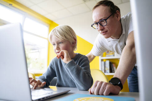 Blond boy using laptop by professor leaning on desk in classroom - NJAF00915