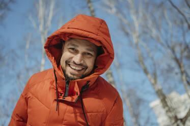Smiling man wearing orange color winter jacket at park - ANAF02748