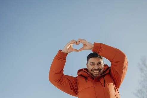Smiling man showing making heart shape gesture with sky in background - ANAF02753