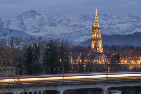 Schweiz, Kanton Bern, Bern, Langzeitbelichtung eines vorbeifahrenden Zuges in der Abenddämmerung mit dem Berner Münster und den Alpen im Hintergrund - KEBF02850