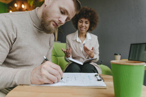 Businessman planning strategy on clipboard sitting by businesswoman in office - YTF01981