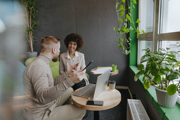 Lächelnde Geschäftsfrau hört einem Geschäftsmann zu, der in einem Coworking-Büro seine Strategie erklärt - YTF01988