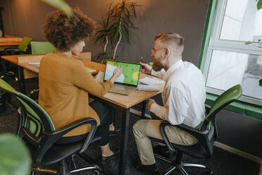 Multiracial business colleagues discussing over laptop sitting at desk in coworking office - YTF02012