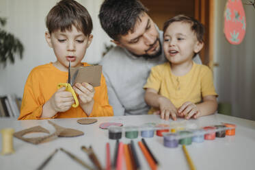 Junge schneidet Karton mit einer Schere in der Nähe von Vater und Bruder zu Hause - ANAF02760