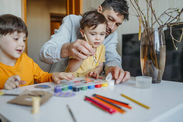 Father with children painting cardboard Easter eggs at table - ANAF02762