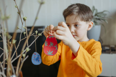 Junge dekoriert Ostereier aus Pappe auf einem Zweig zu Hause - ANAF02770