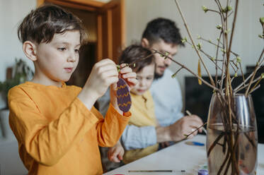 Boy decorating cardboard Easter egg on twig with father and brother in background - ANAF02772