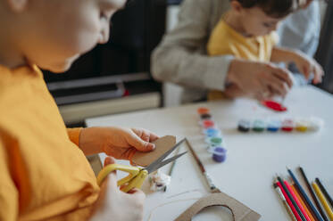 Junge schneidet Karton mit einer Schere in der Nähe von Vater und Bruder, die im Hintergrund malen - ANAF02775