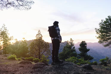 Mann mit Rucksack steht auf einem Felsen in der Nähe von Bäumen bei Sonnenuntergang - ASGF04940