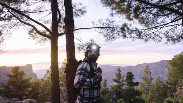 Man wearing headlamp and standing near trees in front of mountains - ASGF04948