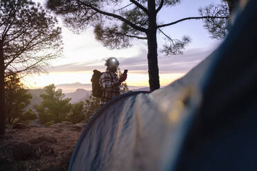 Man wearing headlamp and holding smart phone near trees at sunset - ASGF04950