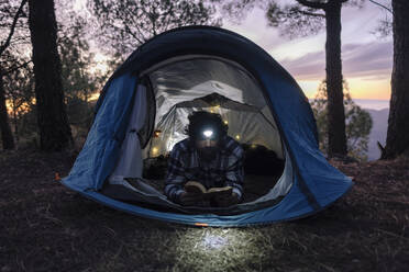 Man wearing headlamp and reading book in tent - ASGF04952