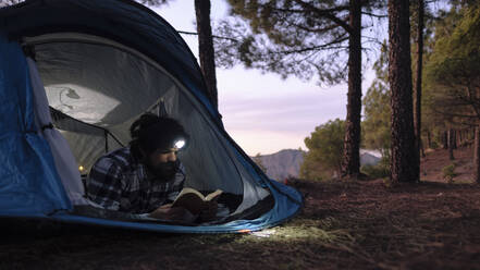 Man wearing headlamp and reading book in tent at sunset - ASGF04953