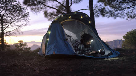 Man wearing headlamp and reading book in tent near trees - ASGF04954