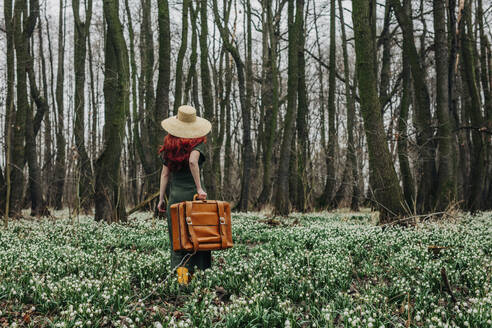 Woman walking with vintage suitcase through flowers in forest - VSNF01743