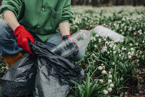 Woman collecting plastic bottle near Lily-of-the-valley flowers in forest - VSNF01747