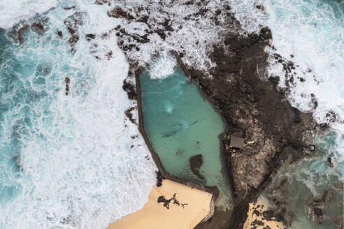 Luftaufnahme einer Drohne von großen Meereswellen, die sich über Felsen brechen und ein Gezeitenbecken mit schwimmenden Kindern überfluten, Boucan Canot, Saint Paul, La Réunion. - AAEF27937