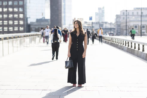 Glückliche junge Frau steht mit Handtasche auf der Straße in London Stadt - WPEF08561