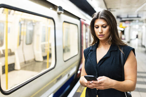 Young woman using smart phone near subway train at platform - WPEF08577