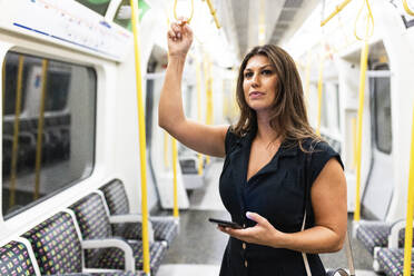 Young woman holding mobile phone and standing in subway train - WPEF08579