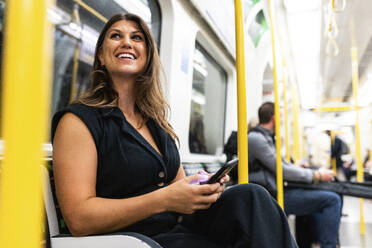 Glückliche junge Frau mit Mobiltelefon im Zug sitzend - WPEF08590