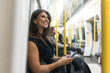 Happy young woman sitting with mobile phone in subway train - WPEF08591