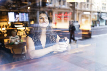 Glückliche junge Frau mit Kaffeetasse sitzend und durch Glas gesehen - WPEF08602